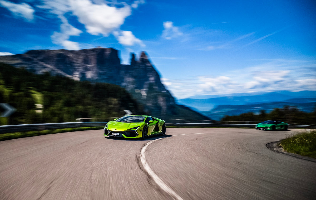 A bright lime green Lamborghini Revuleto driving up a winding mountain road surrounded by mountain peaks, followed by a darker green Lamborghini Temerario